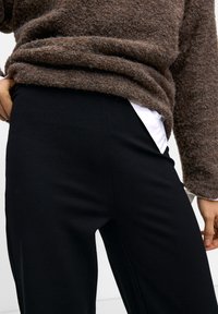 Brown textured sweater with loose fit, worn over black fitted pants with a smooth surface. White shirt hem visible beneath the sweater.