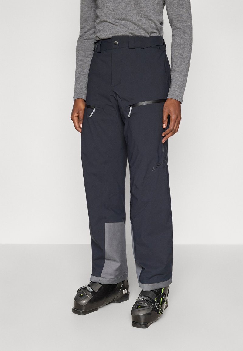 Αδιάβροχο, σκούρο μπλε παντελόνι σκι με ενισχυμένο γκρι μπαλώμα, τσέπες με φερμουάρ και ρυθμιζόμενο ζωνάρι μέσης, σχεδιασμένο για απόδοση σε κρύο καιρό.
