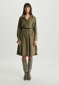 Robede-shirt vert olive avec une taille ceinturée et des manches longues, associée à des bottes montantes en imprimé serpent. Tissu lisse, design fluide.