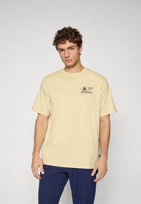 Light yellow cotton t-shirt with a crew neck and short sleeves. Features playful graphic print on the left chest area.