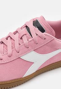 Sapatilha de camurça rosa com logotipo em couro branco, apresentando uma superfície texturizada, atacadores redondos e uma sola de borracha castanha.