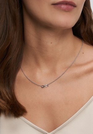 Sølvkæde med et uendelighedssymbol. Kæden er delikat, med en glat tekstur, båret på bar hud.