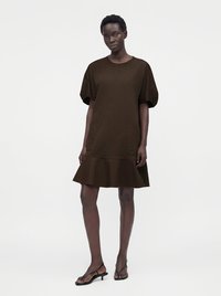 Femme debout portant une robe marron foncé à manches courtes avec un ourlet évasé et des sandales noires à talons bas sur un fond uni.