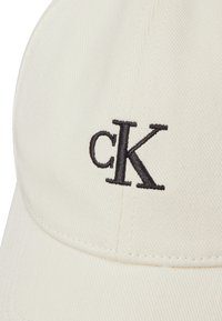 White baseball cap featuring a black embroidered "cK" logo. Made from textured fabric with a curved brim and stitched detailing.