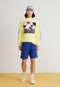 Κίτρινο μακρυμάνικο πουκάμισο με γραφικό σχέδιο, μπλε σορτς και λευκά αθλητικά παπούτσια; διαθέτει καρό σχέδιο στα μανίκια και ελαφριά υφή υφάσματος.