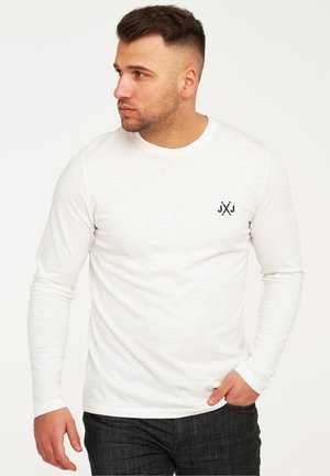 Weißes Langarm-T-Shirt aus weichem Material, mit Rundhalsausschnitt und einem kleinen schwarzen Logo auf der linken Brust. Kombiniert mit dunklen Jeans.