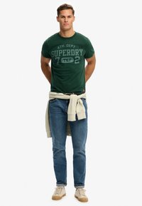 Zielona bawełniana koszulka z nadrukowanym logo, zestawiona z niebieskimi dżinsami i beżowymi trampkami. Lekki sweter związany w talii.