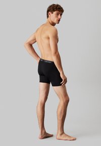 Crne pamučne bokserice s uskim dizajnom i elastičnom trakom u kojoj se nalazi bijeli logo "Calvin Klein". Model stoji bos, okrenut bokom.