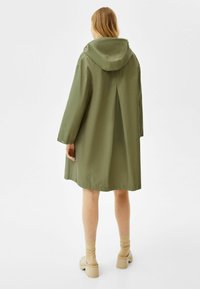 Green hooded coat with a relaxed fit, featuring a back pleat, long sleeves, and button details at the cuffs. Smooth fabric texture.