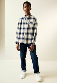 Flanellhemd in Creme, Marine und Blau kariert mit Brusttasche; kombiniert mit dunkelblauen Jeans und weißen Sneakern. Glatte Stofftextur.