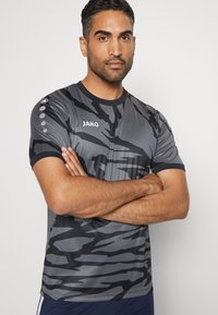 Grijs sportshirt met korte mouwen en zwart camouflagepatroon, ronde hals en zwarte accenten. "JAKO" logo op de voorkant, cirkelvormig detail op de schouder.