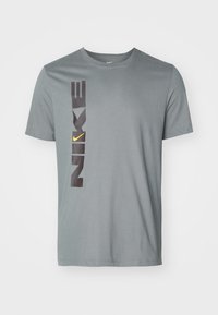 Grå bomulls-t-shirt med stort vertikalt "NIKE"-tryck i svart och en gul swoosh-accent på vänster sida, rund halsringning, korta ärmar.