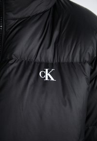 Svart boblejakke med en glatt, blank overflate og en hvit "cK"-logo på venstre bryst. Har høy krage og glidelås.
