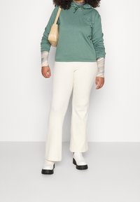 Grön huvtröja med en framlogo, parat med krämfärgade utsvängda byxor och vita ankelstövlar. Modellen håller en beige axelväska.