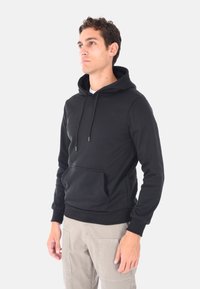 Uomo che indossa un hoodie nero con tasca frontale e pantaloni cargo beige, in piedi e che guarda di lato su uno sfondo neutro.