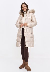 Puffer coat in shiny beige with a faux fur hood, quilted design, front pockets, and zipper closure. Worn over a cream sweater and brown pants.