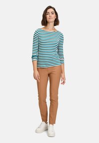 Blå och beige randig långärmad skjorta kombinerad med karamellfärgade slim-fit byxor och vita sneakers, stående mot en enfärgad bakgrund.