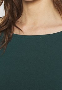 Mörkgrön torso-plagg med bred halsringning, tillverkat av ett slätt, stretchigt material. Har inga synliga mönster eller utsmyckningar.