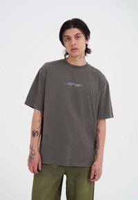 Šedé volné tričko vyrobené z bavlny s kulatým výstřihem, s malou bíle vyšitou logem na hrudi a viditelnými detaily šití.