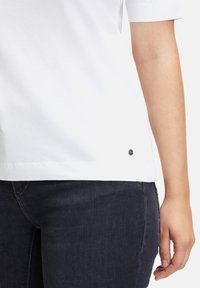 Biała bawełniana koszulka z krótkim rękawem, z okrągłym dekoltem i małym czarnym logo na dolnym brzegu. Zestawiona z ciemnoniebieskimi dżinsami.