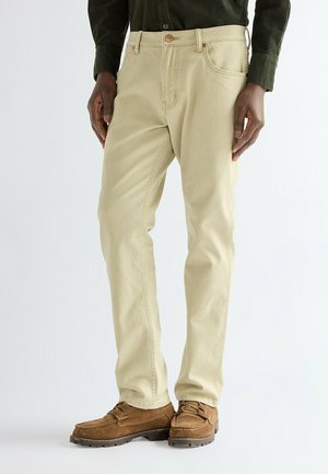 Uomo che indossa pantaloni beige, scarpe in camoscio marrone e una camicia verde scuro con bottoni, in piedi su uno sfondo bianco semplice.