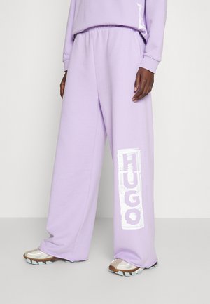 Osoba nosząca luźne lawendowe spodnie dresowe z białym logo "HUGO" na lewej nodze oraz beżowe buty sportowe z grubymi podeszwami.