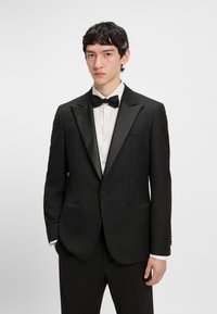 Noor mees, kes kannab musta satiinist kraega smokki, valget kleidisärki ja musta kikilipsu, seistes tavalise heleda tausta ees.