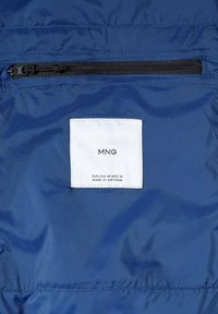 Navy sininen takin sisustus, jossa on valkoinen kangaslabeli, jossa lukee "MNG" ja hoito-ohjeet. Sisältää mustan vetoketjullisen taskun labelin yläpuolella.