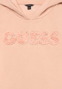 Pērļu krāsas kapuce ar vārdu "GUESS", kas izveidota no teksturētiem auduma ziediem, ko apvilkuši mazi sudraba pērles uz krūtīm.