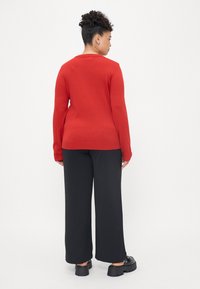 Sweater de malha vermelho de mangas longas com decote redondo e punhos canelados, combinado com calças largas pretas e sapatos pretos.