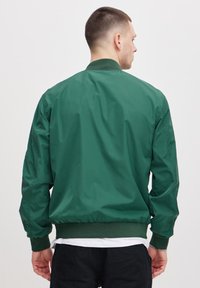 Grønn bomberjakke med en glatt tekstur, ribbestrikkede mansjetter og krage. Har et enkelt design, elastisk liv og ingen synlige mønstre.