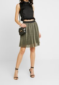 Crop top en satin noir avec bordure en plumes, associé à une jupe métallique vert olive. Un sac bandoulière noir et des talons noirs à lanières complètent le look.