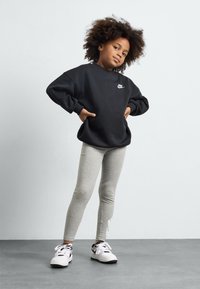 Fekete kapucnis pulóver fehér Nike logóval, szürke leggings és fehér sportcipő fekete akcentussal. A gyermek magabiztosan áll, kezét a csípőjére téve.