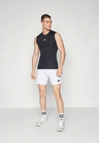 Zwart mouwloos sporttopje met een wit logo, gecombineerd met witte shorts met een zwart logo. Het model draagt witte sneakers en gestreepte sokken.