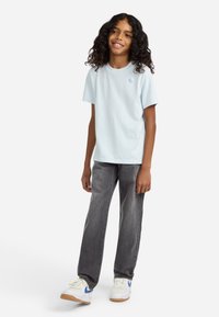 Niño con cabello rizado, vistiendo una camiseta de color azul claro, pantalones vaqueros oscuros y zapatillas blancas con acentos azules, de pie y sonriendo.