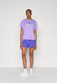 Μωβ γραφικό t-shirt με εκτύπωση βουνού, συνδυασμένο με μπλε σορτς με υφή. Τα παπούτσια είναι λευκά με ανοιχτό γκρι λεπτομέρειες. Μινιμαλιστικός σχεδιασμός.