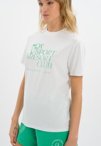 Biała bawełniana koszulka z zielonym napisem "SPORT & RESORT CLUB". Okrągły dekolt, krótkie rękawy, luźny krój. Połączona z zielonymi szortami.