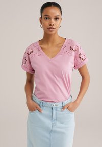 Tricou din bumbac roz cu guler în V, decorate cu broderie florală din dantelă pe mâneci. Asortat cu o fustă din denim albastru deschis.