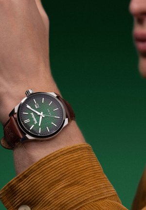 Hodinky so zeleným vzorovaným ciferníkom, strieborným púzdrom a hnedým koženým remienkom, nosené na zápästí. Pozadie je zelené, odhaľujúce textúru látky košele.