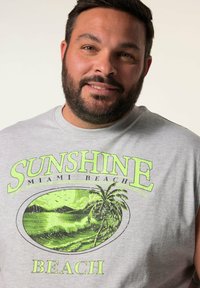 Maglietta senza maniche grigia con una vivace grafica verde di palme e onde, con il testo "SUNSHINE MIAMI BEACH" in evidenza.