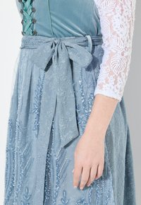 Lichtblauwe jurk met kant en met kralen versierde bloemenpatronen, met een stoffen ceintuur die in de taille is gestrikt. Witte kanten mouwen voegen textuur toe.