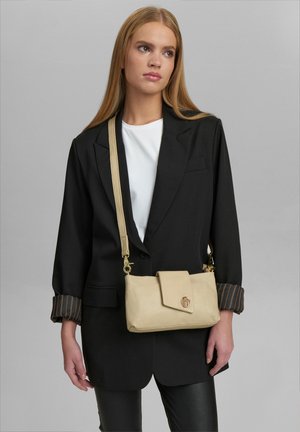 Borsa a tracolla in pelle beige con chiusura a pattina e logo in evidenza, dotata di una lunga tracolla e superficie liscia, tenuta da una persona in un blazer nero.