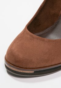 Chaussure fermée en daim marron avec un devant arrondi, dotée d'un petit talon et d'une semelle noire lisse. Surface texturée avec un léger éclat.