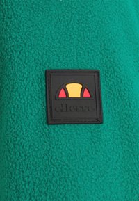 Tessuto in pile verde con una toppa in gomma nera che presenta un design giallo e rosso. La superficie strutturata aggiunge profondità al materiale.