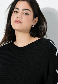 Junge Frau mit langen dunklen Haaren, die silberne Ohrringe und einen schwarzen Strickpullover mit weißen Details trägt, leicht nach links gewandt vor einem weißen Hintergrund.