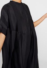 Kvinna i en lös svart klänning med vertikala ränder och samlad midja, arm utsträckt åt sidan mot en enkel bakgrund.