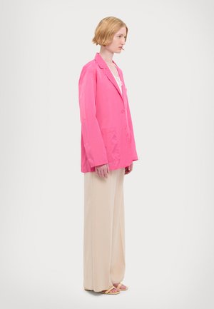 Rosa übergroßer Blazer mit glatter Textur, zwei Vorderaschen und einem einreihigen Knopfverschluss, kombiniert mit beigen Hosen und gelben Sandalen.
