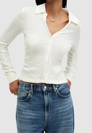 Femme portant un haut blanc texturé à manches longues boutonné et un jean bleu en denim, les mains posées sur les hanches et un collier en perles dorées.