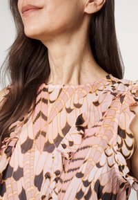 Vaaleanpunaiseen kukkakuosisessa paidassa on röyhelöiset hihat. Suunnittelussa on tumma ruskea ja kermaelementtien kuvioita kevyessä kankaassa.