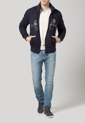 Chaqueta de navy con cremallera y cuello alto, bolsillos frontales ajustables y texto gráfico en blanco. Lleva puesta sobre una camiseta de color beige claro y jeans de color azul claro.